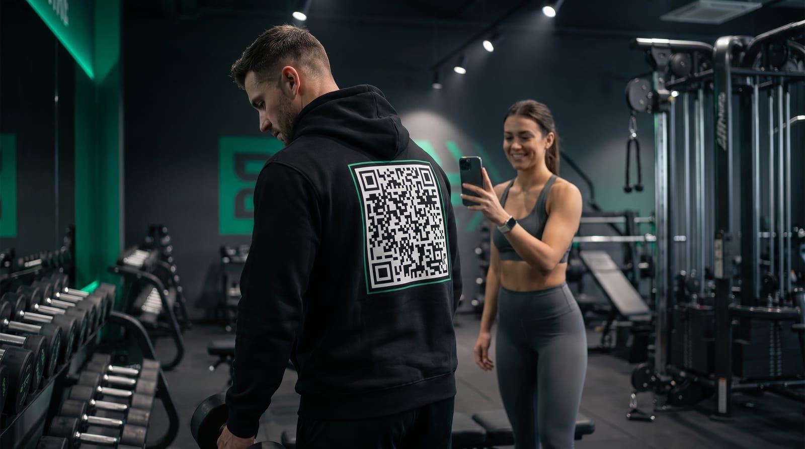 Person im Fitnessstudio trägt einen Hoodie mit QR-Code auf dem Rücken, eine andere Person scannt ihn